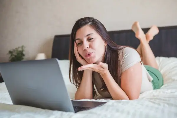 Lächelnde Frau auf dem Bett liegend flirtet während Nacktchat auf Laptop - Sexchat und Sexdate Tipps